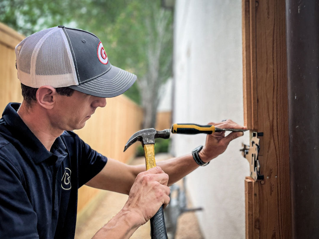 A handyman in a cap and polo shirt using a hammer to work on a door frame.