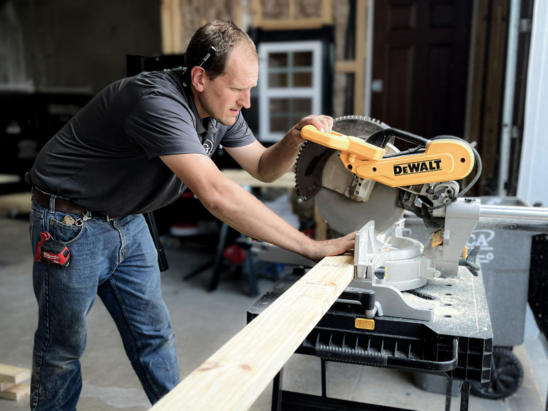 Goodsmith carpenter using a DeWalt miter saw to cut a wooden plank in a workshop.