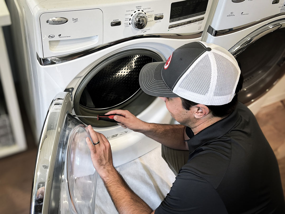 A Goodsmith technician in a baseball cap repairing or examining the inside of a front-loading washing machine, representing a appliance repair service