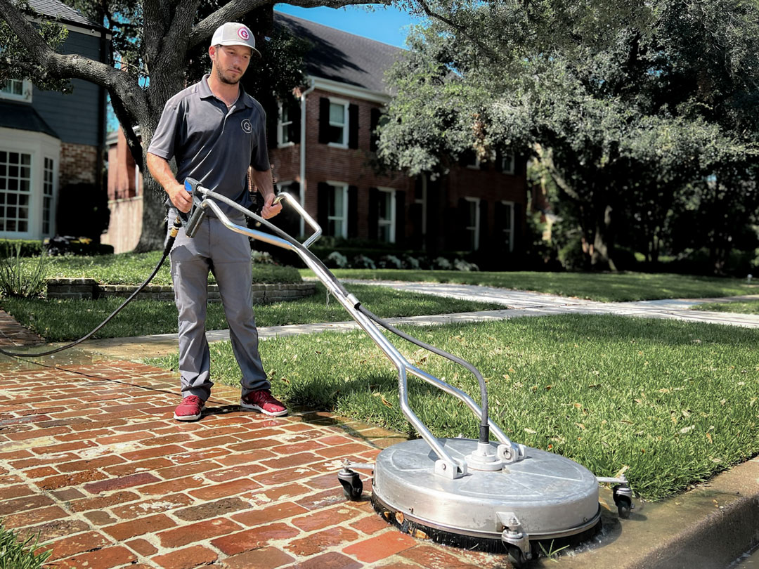 A person using a flat surface cleaner on a brick driveway in front of a house.